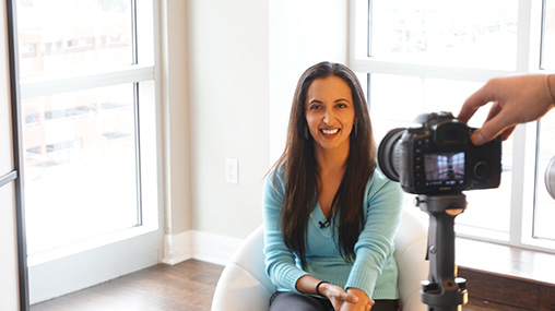 Paula Pant being filmed for the 'Your First Rental Property' course, smiling while seated in a bright, modern space with a camera visible in the frame, representing the professional educational content offered through Afford Anything.
